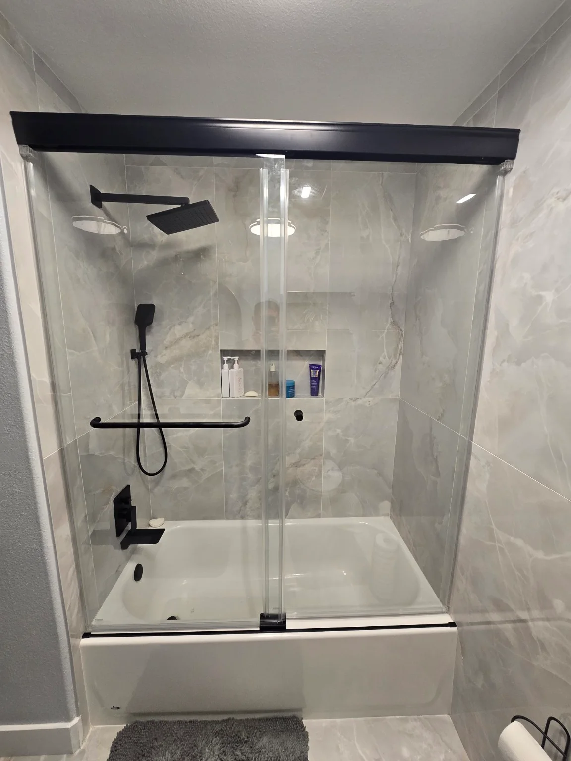 Gray-tiled bathtub opening before glazing and after installation of a black-header sliding glass enclosure — Completed enclosure