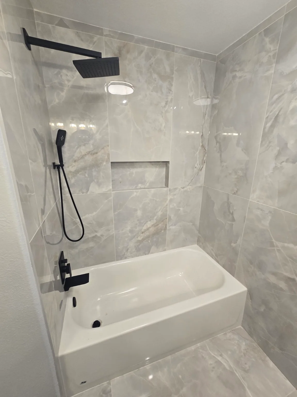 Gray-tiled bathtub opening before glazing and after installation of a black-header sliding glass enclosure — Before glass installation