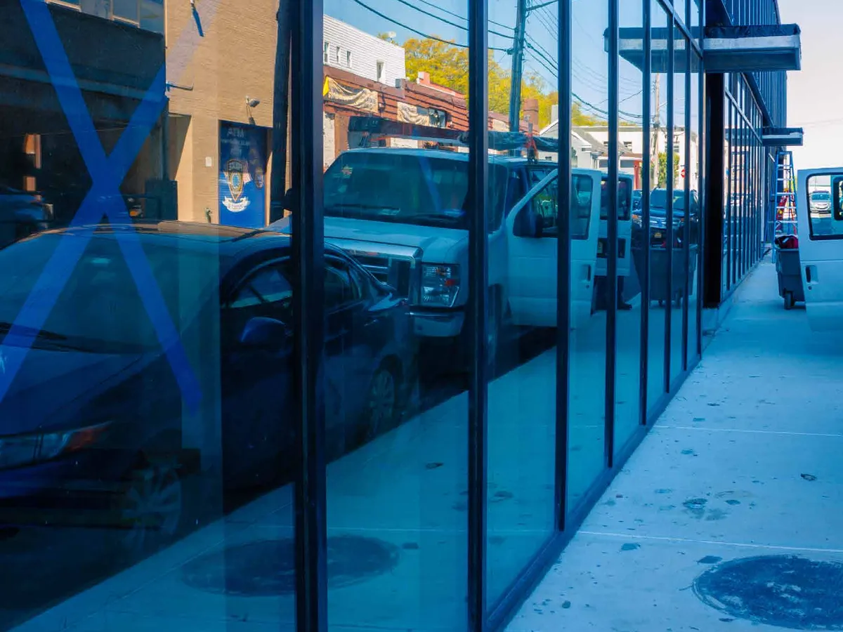 Sidewalk view along a shop's full-height glazed aluminum storefront wall reflecting the street and parked cars.